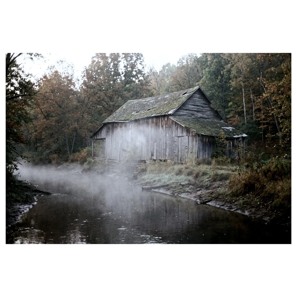 An abandoned wooden sawmill with a sagging roof on a misty riverbank surrounded by autumn trees.